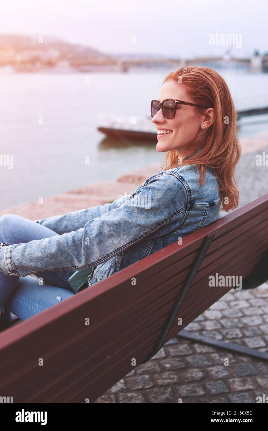 Young balanced redhead Caucasian woman sitting on bench at riverbank side view in sunset Stock Photo