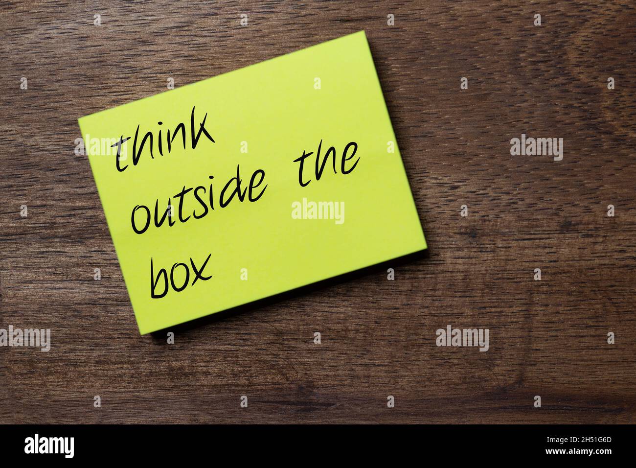 yellow self-adhesive note sticker diagonally on a dark wooden table top with stylized handwriting with the slogan think out of the box Stock Photo