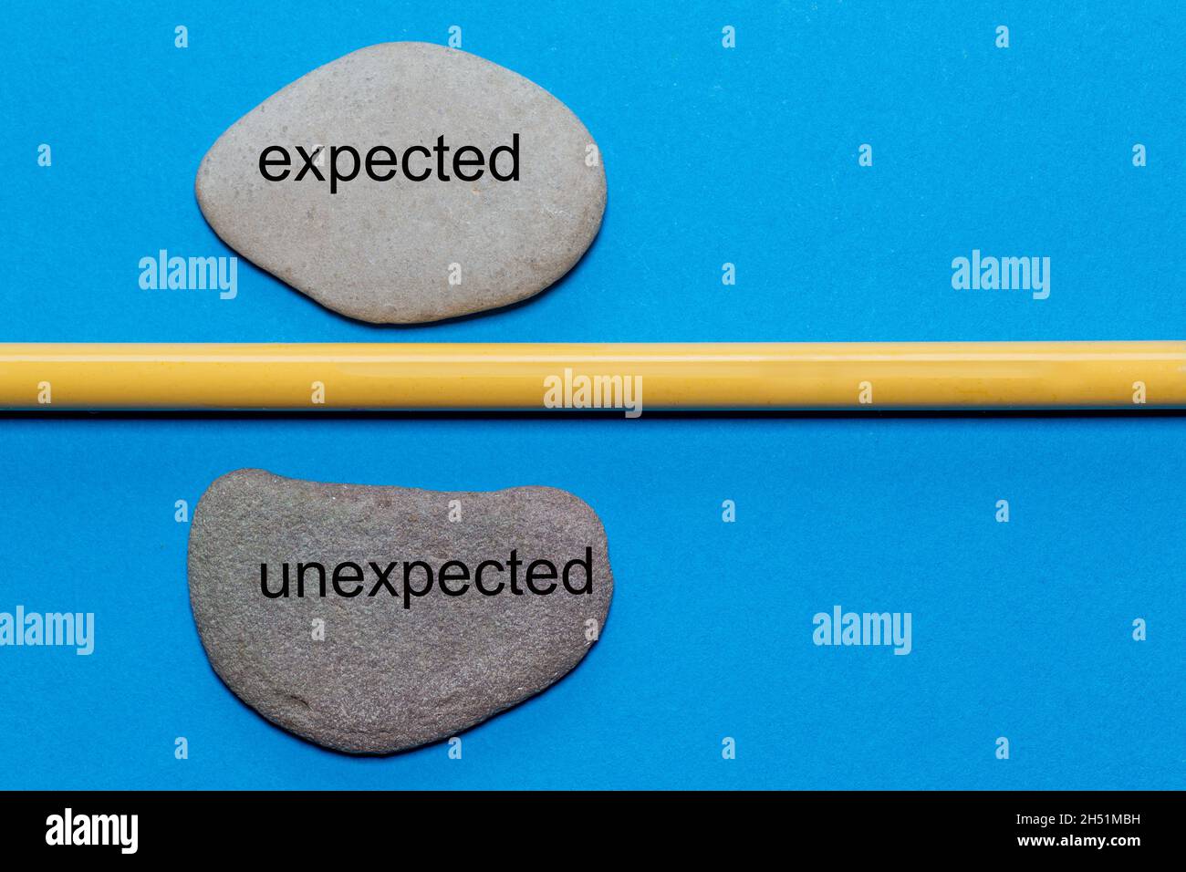 The words expected and unexpected are written on natural smooth stones separated by a yellow pencil. The background is isolated in blue and has a lot Stock Photo