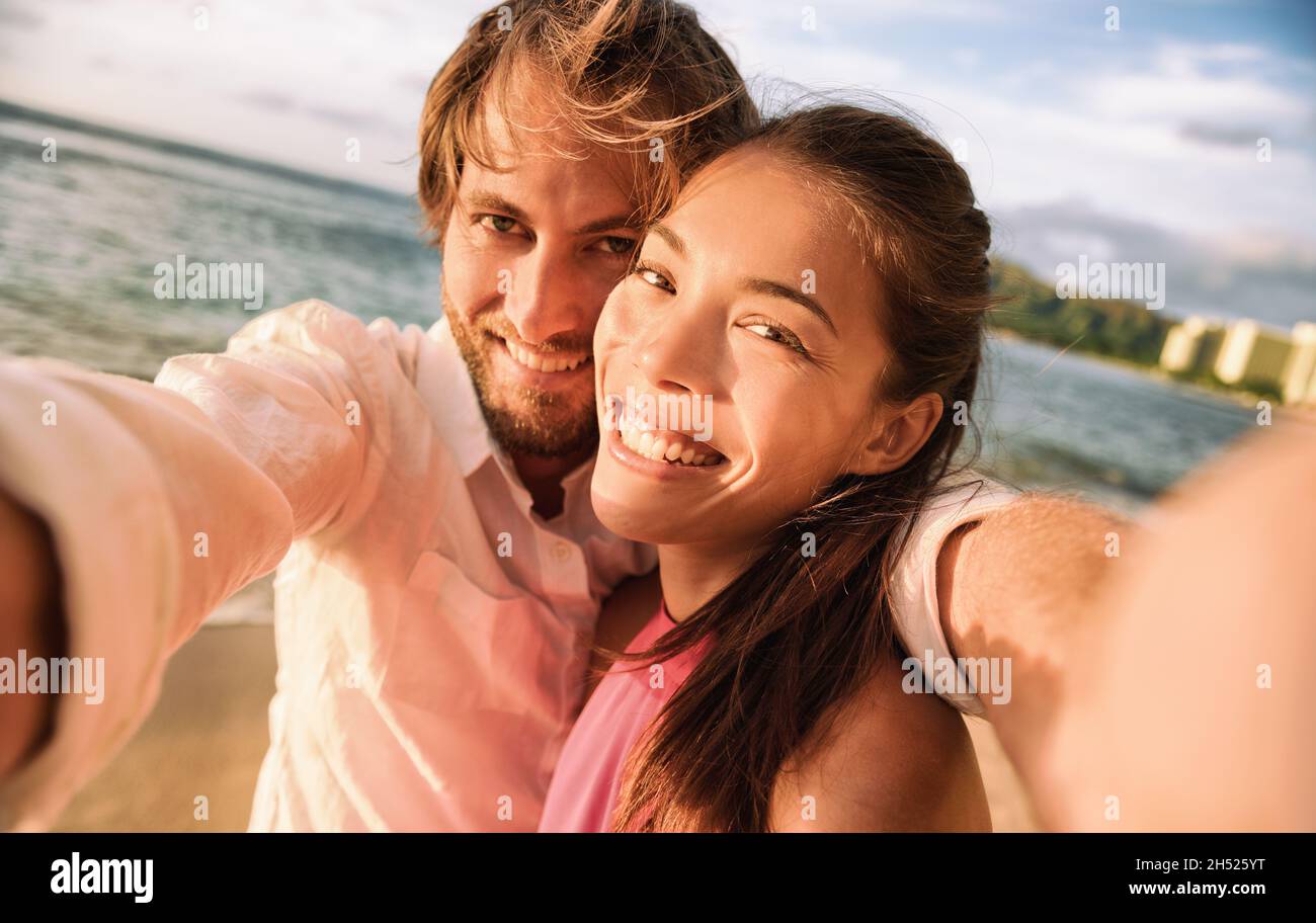 Happy smiling biracial couple taking selfie photo with camera phone. Caucasian man holding smartphone on beach vacation holiday sunset. Stock Photo