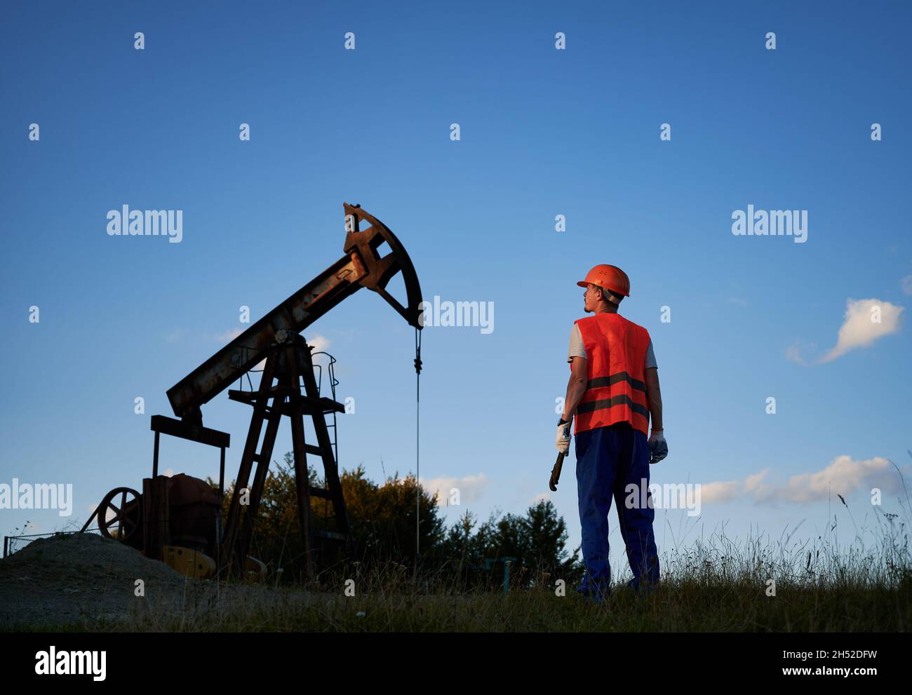 Back view of male worker in protective helmet standing near petroleum pump jack under blue sky. Petroleum engineer in work vest holding industrial wrench and looking at oil pump rocker-machine. Stock Photo
