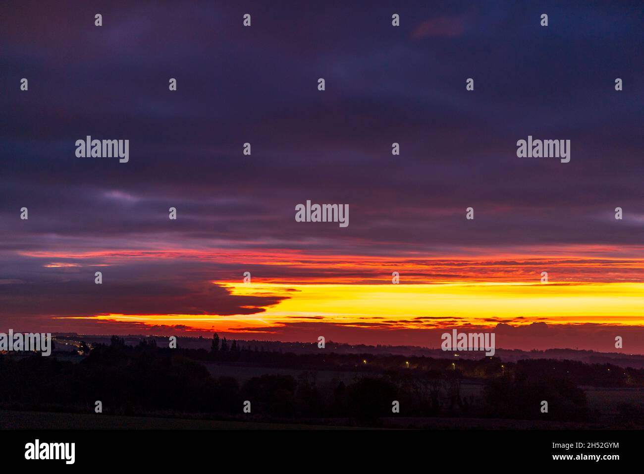 Dark unbroken layer of clouds overhead with a patch of bright yellow and orange cloud on the horizon where dawn's first light breaks through over a silhouetted Kent countryside. Stock Photo