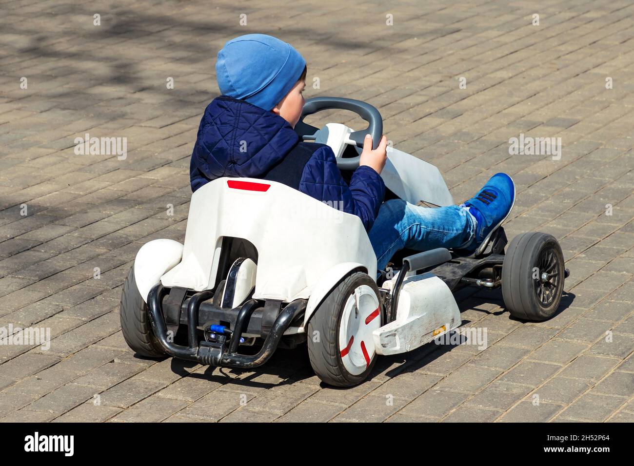 Kid boy back view blue jacket, hat, jeans driving white children pedal go-kart in park springtime. Stock Photo