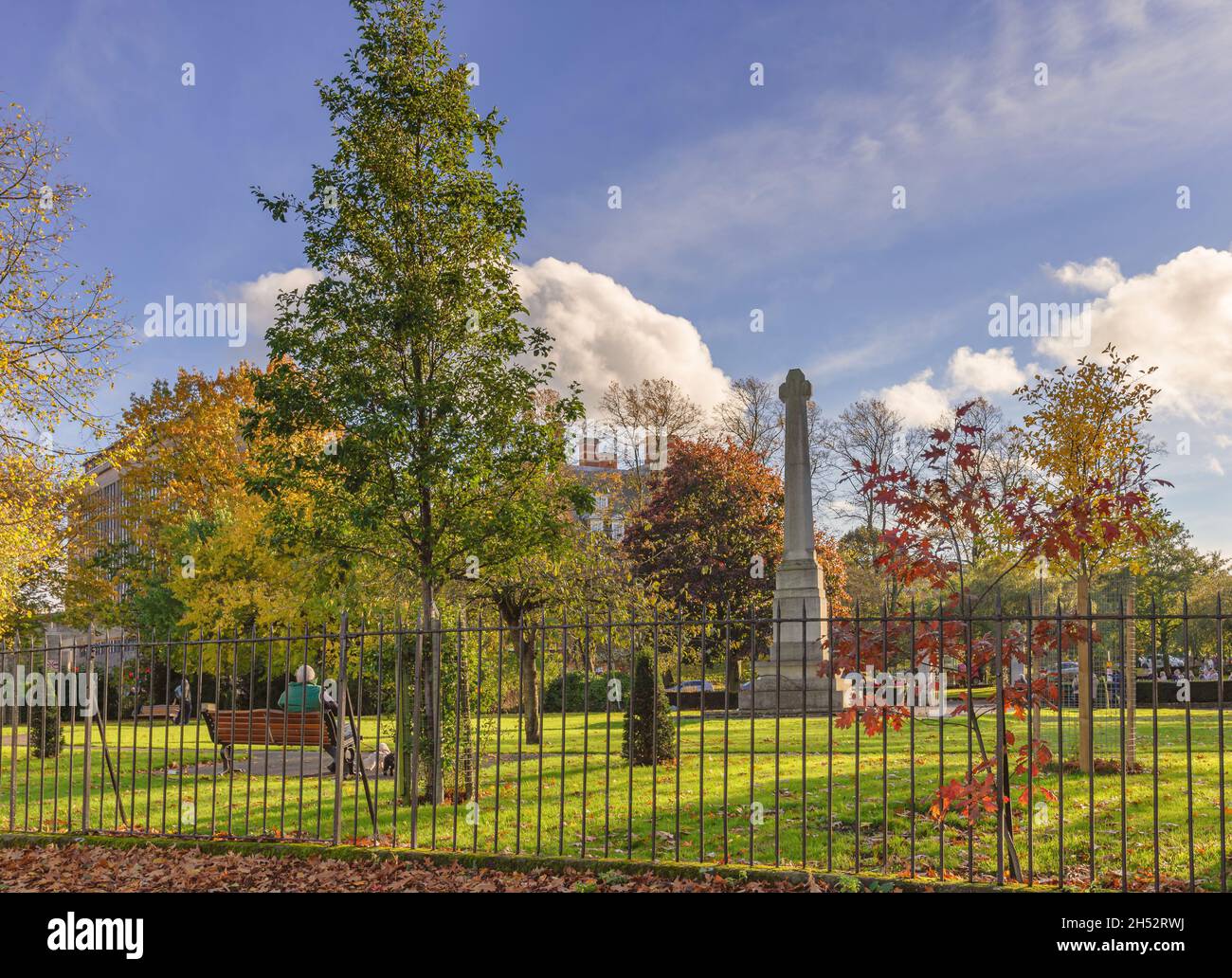 A public garden for remembrance in Autumn. A man sits on a bench and a cenotaph is in the centre of the park. A sky with cloud is above. Stock Photo