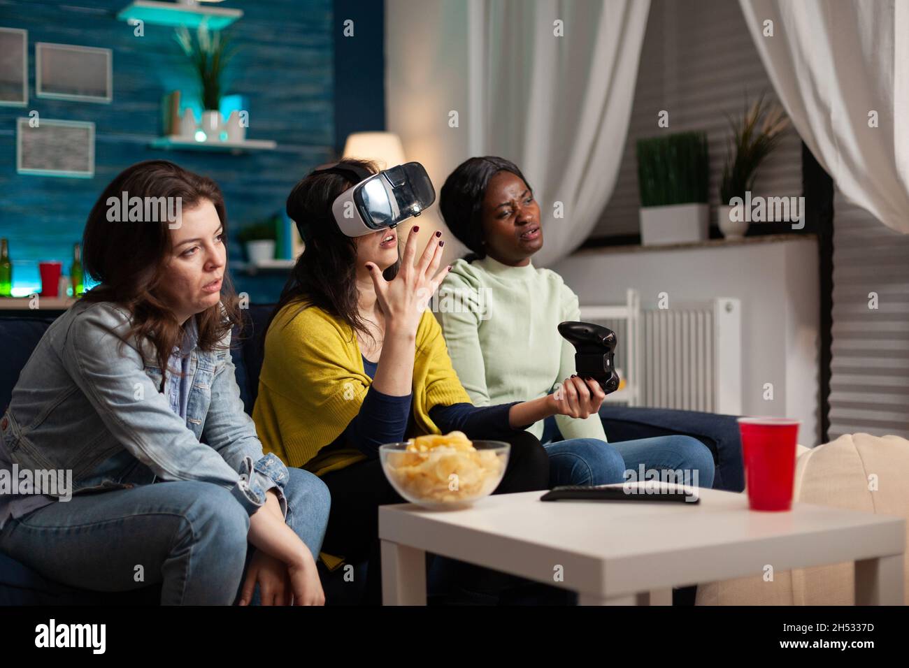 Sad nervous women friends sitting on couh playing online videogames using vr headset and joystick losing online competition. Multiethnic people enjoying hanging out together making fun activity Stock Photo