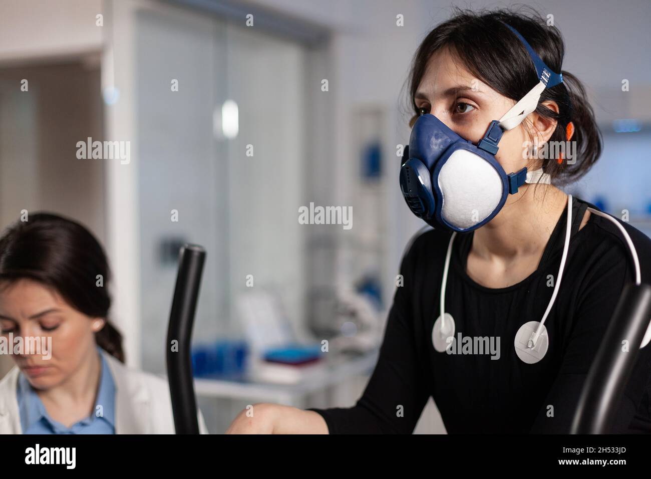 Athlete woman with mask running on fitness bycle trainning body endurance while specialist doctor measuring heart rate monitoring EGK data in laboratory. Sportwoman with medical electrodes on it Stock Photo
