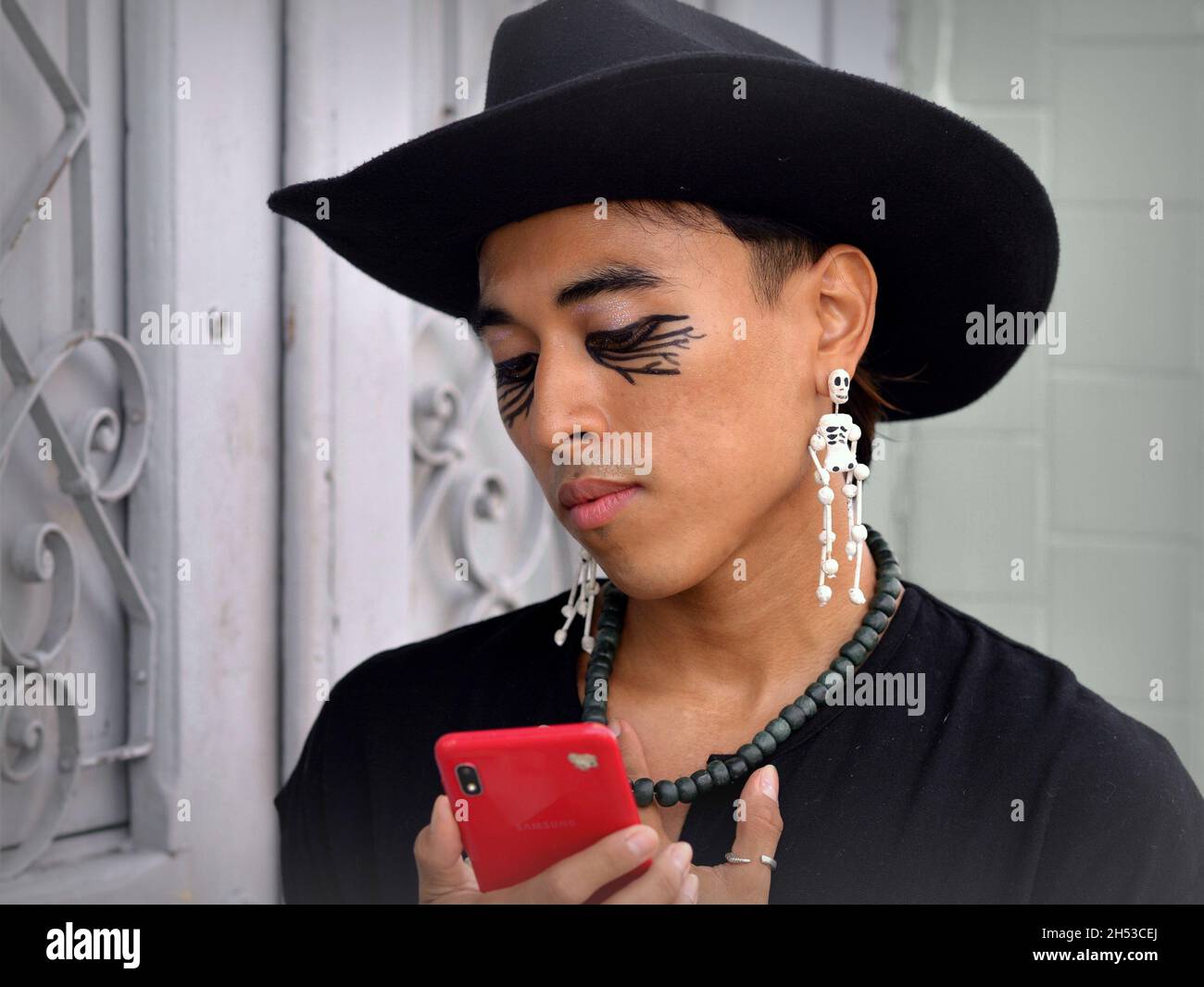 Handsome young nonbinary Mexican Latino with Day of the Dead skeleton earrings wears a black hat and looks at his red smart phone. Stock Photo