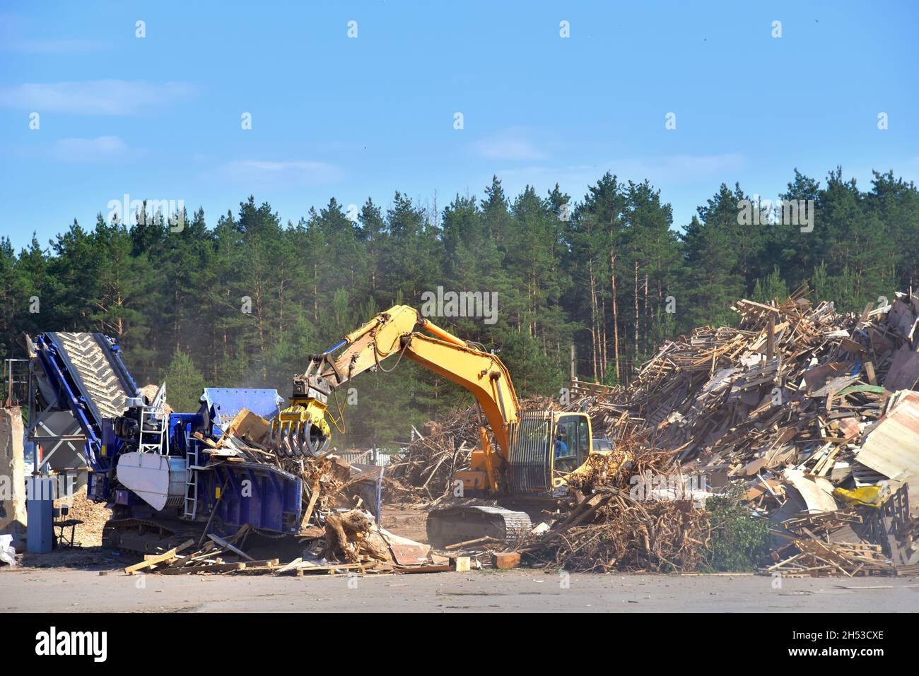 Excavator with log grab crane loads round logs in woodchipper. Mobile chipper for wood chipping. Grab loads timber for lumber mill. Horizontal Grinder Stock Photo