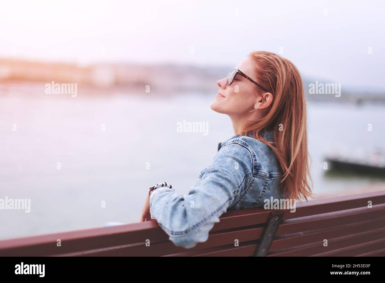 Young balanced redhead Caucasian woman sitting on bench at riverside meditating in sunset Stock Photo