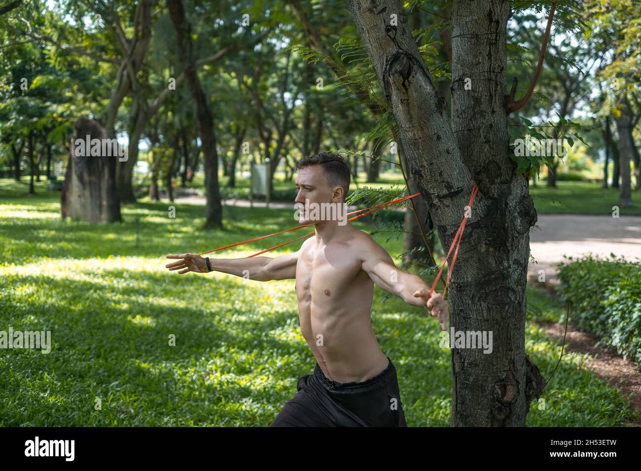 Sporty active young man in black sportswear doing sport exercises in the park. Muscular fit man doing exercises with sports elastic band outdoors. Stock Photo