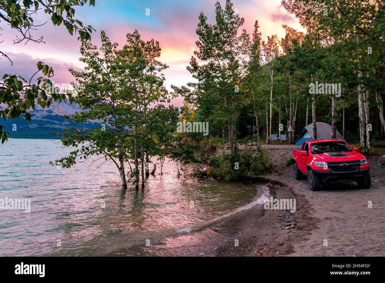 The Perfect camping spot on Abraham Lake with trees, tent and truck infront of sunset lit sky with mountains in background Stock Photo