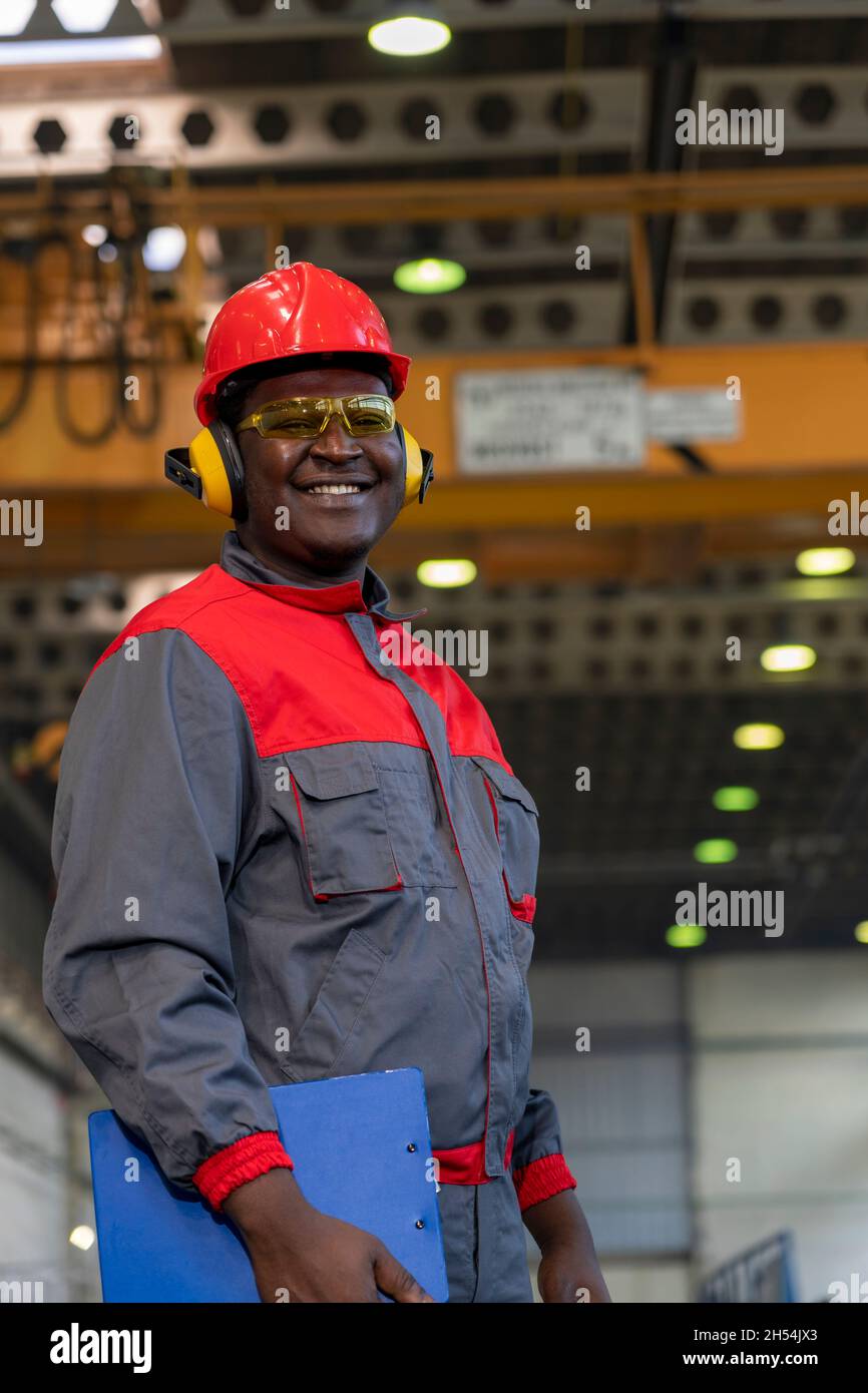 Waist Up Portrait Of Black Industrial Worker In Red Helmet, Yellow Safety Goggles, Noise Reduction Earmuffs And Work Uniform In A Factory. Stock Photo