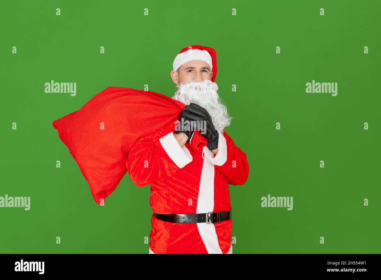 A Caucasian man dressed as Santa Claus is holding a red sack closed over his shoulder. The background is green. Stock Photo