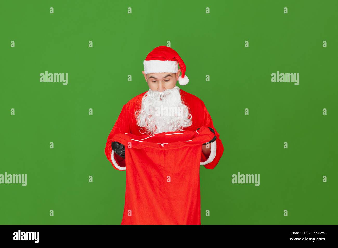 A Caucasian man dressed as Santa Claus is holding a red cloth sack open while looking inside with a surprised expression. The background is green. Stock Photo