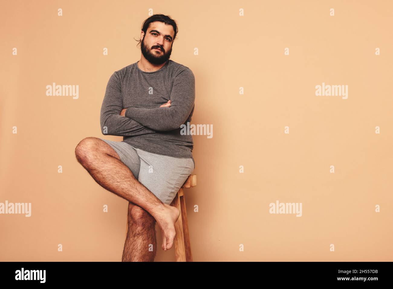 Confident young man sitting with his arms crossed on a wooden chair. Body positive young man looking at the camera in a studio. Self-assured young man Stock Photo