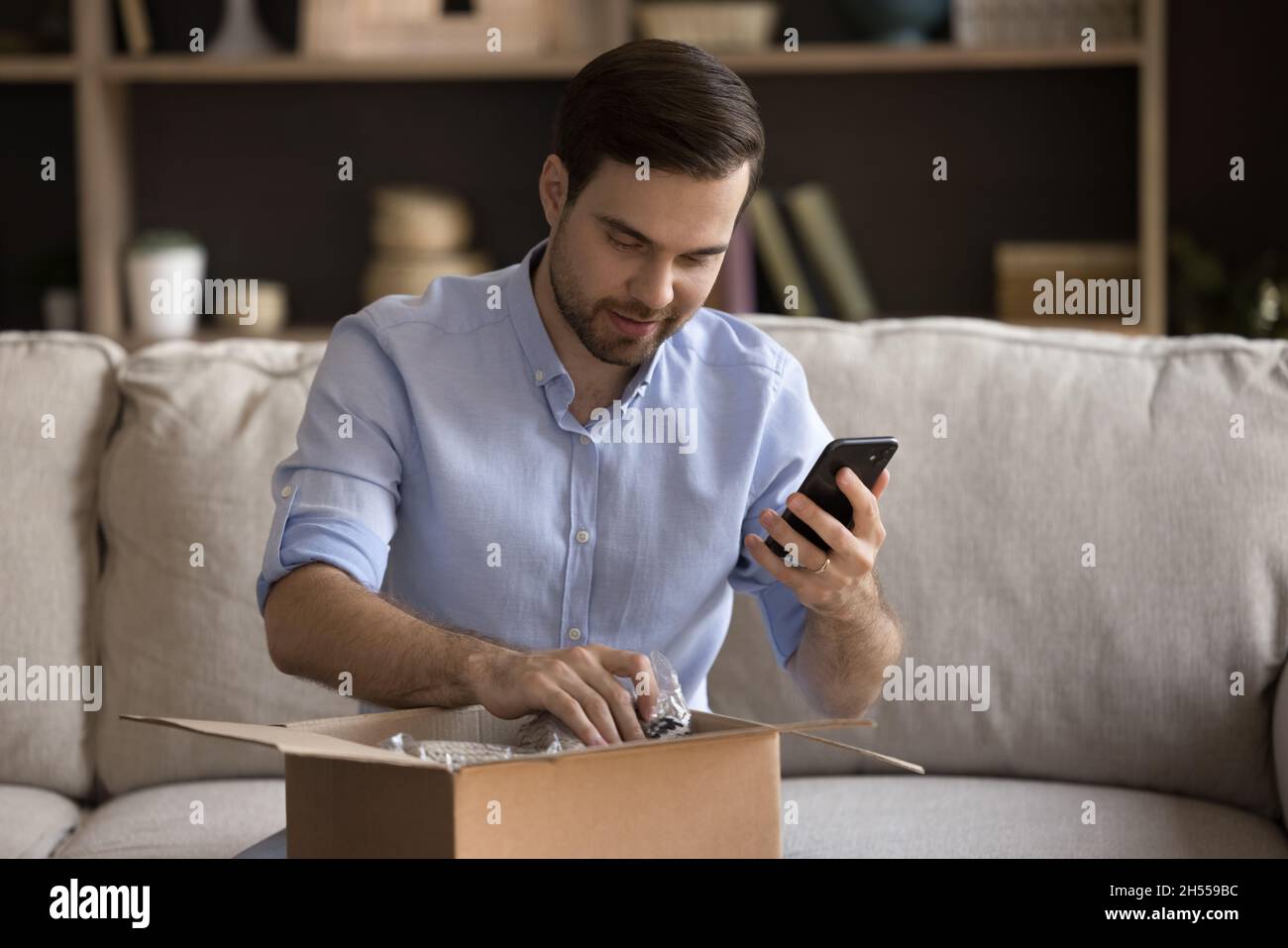 Happy young man unpacking parcel, leaving review in mobile app. Stock Photo