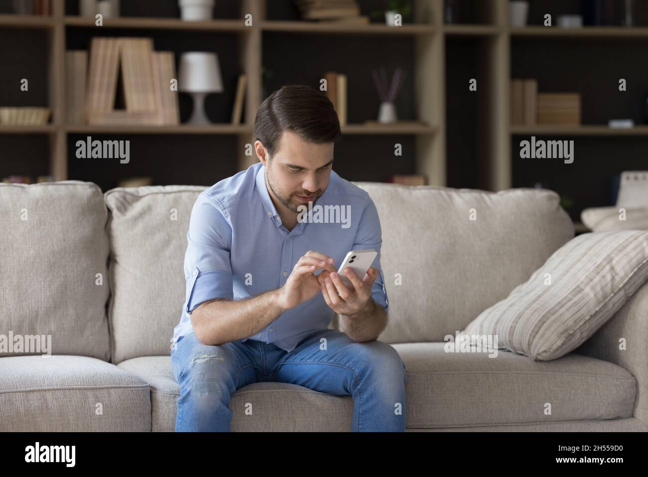 Happy millennial man using smartphone, sitting on sofa. Stock Photo