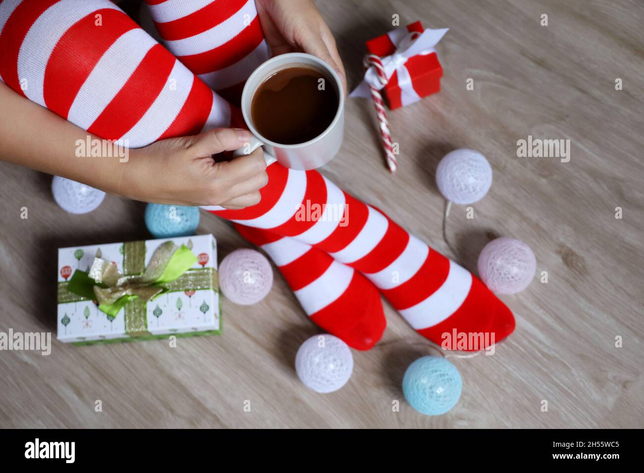 Female legs in Christmas knee socks on a floor near the gift boxes and toy balls. Woman sitting with cup of hot cocoa in hands, New Year celebration Stock Photo
