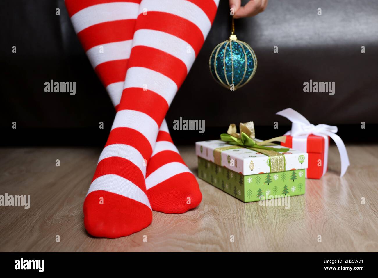 Female legs in Christmas knee socks on a floor near the gift boxes. Woman sitting on a bed with toy ball in hand, New Year celebration Stock Photo
