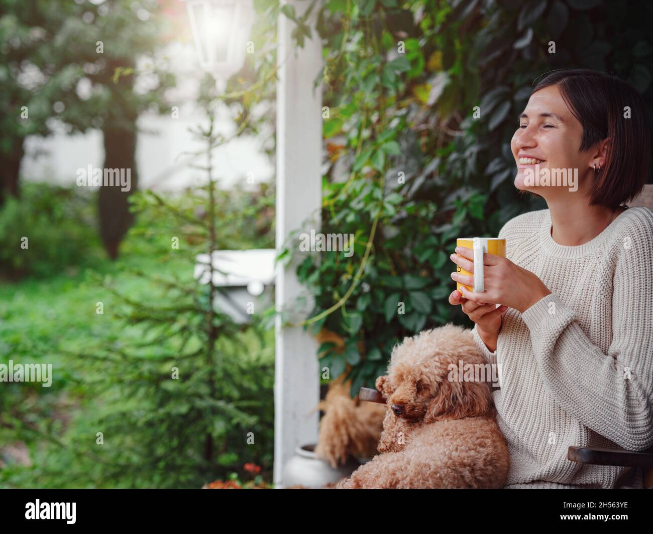 Young asian woman resting outdoors with cup of coffee on porch of country house, chilling outside with poodle dog . autumn lifestyle, leisure free time concept. Copy space Stock Photo