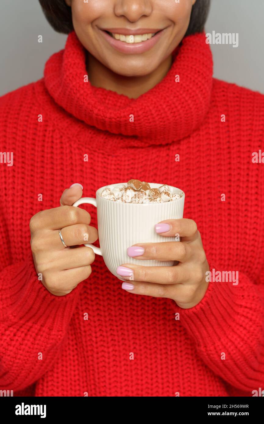 Happy smiling african female in cozy red sweater hold cup of cocoa or hot chocolate with marshmallow Stock Photo