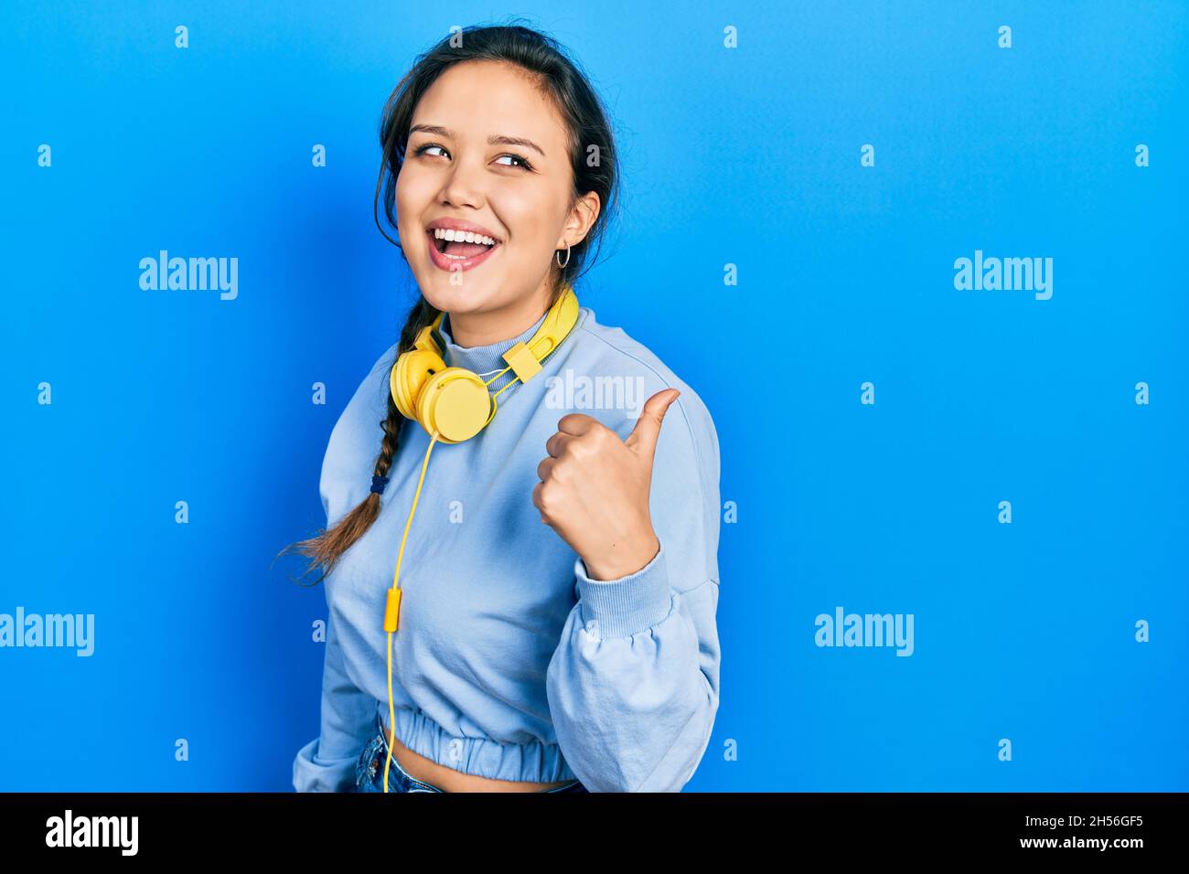 Young hispanic girl listening to music using headphones pointing thumb ...