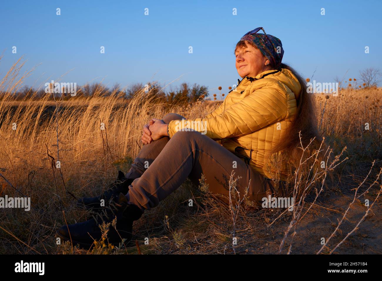 A pretty happy blonde with long hair sits on the ground and watches the sun set. Stock Photo