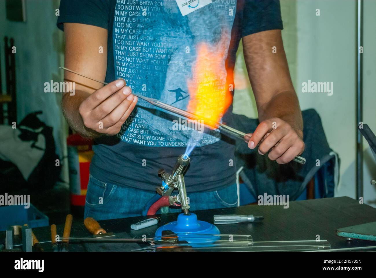 A young man holds a glass pipe into the flame of a  burner during a glassblowing workshop on the MBO (secondary vocational education)  info market Stock Photo