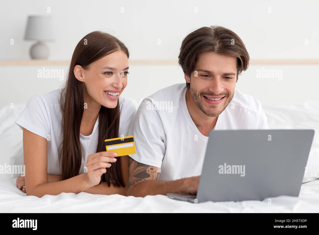 Happy millennial caucasian woman and guy lie on bed paying for purchase, order with laptop and credit card Stock Photo