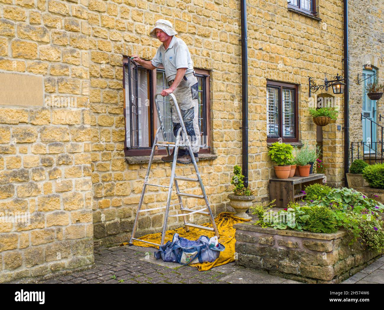 A man in overalls standing on a ladder while working on a window frame at the front of his house. Stock Photo
