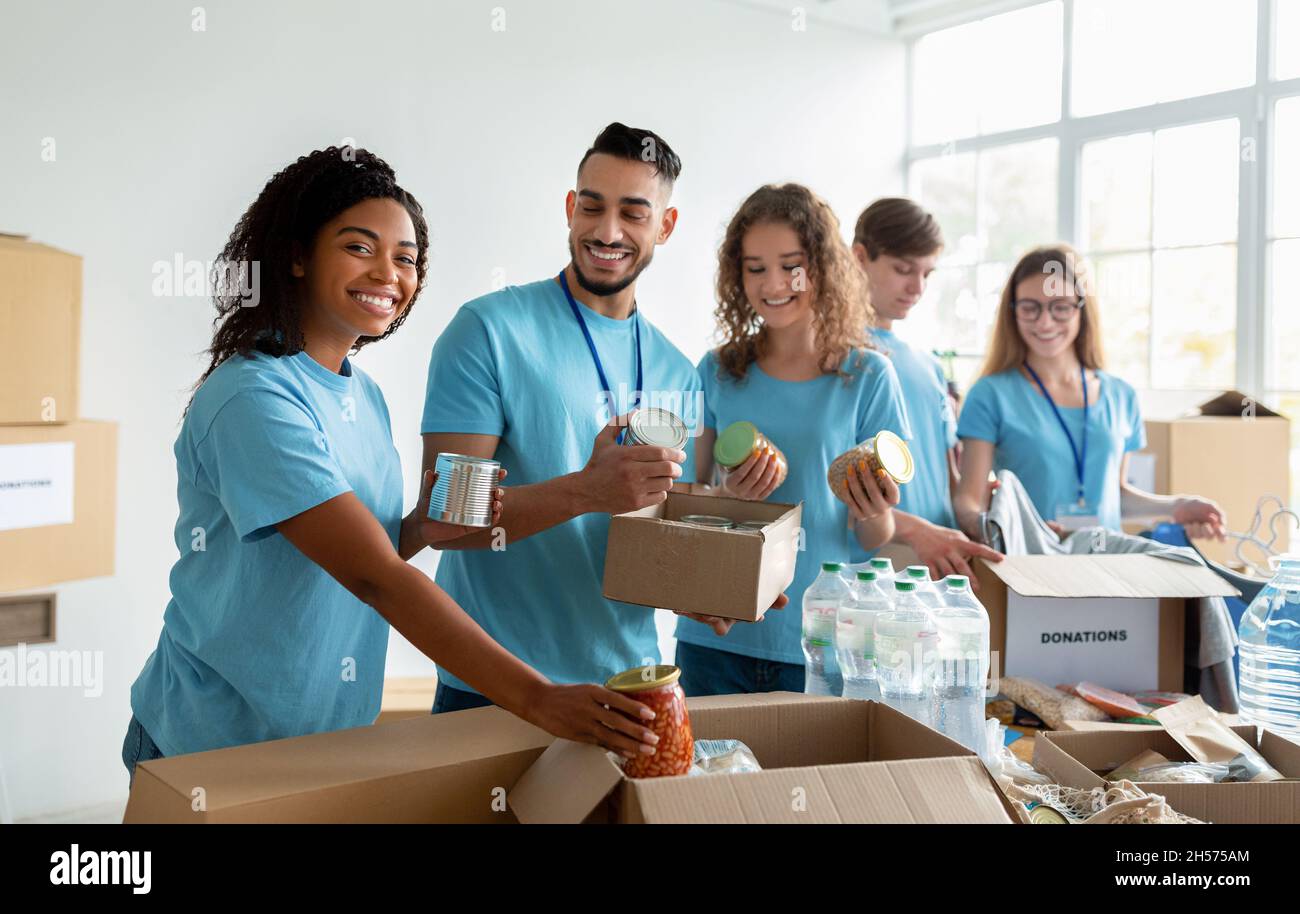 Diverse group of volunteers sorting donated food in boxes, working in community charity donation center and smiling Stock Photo