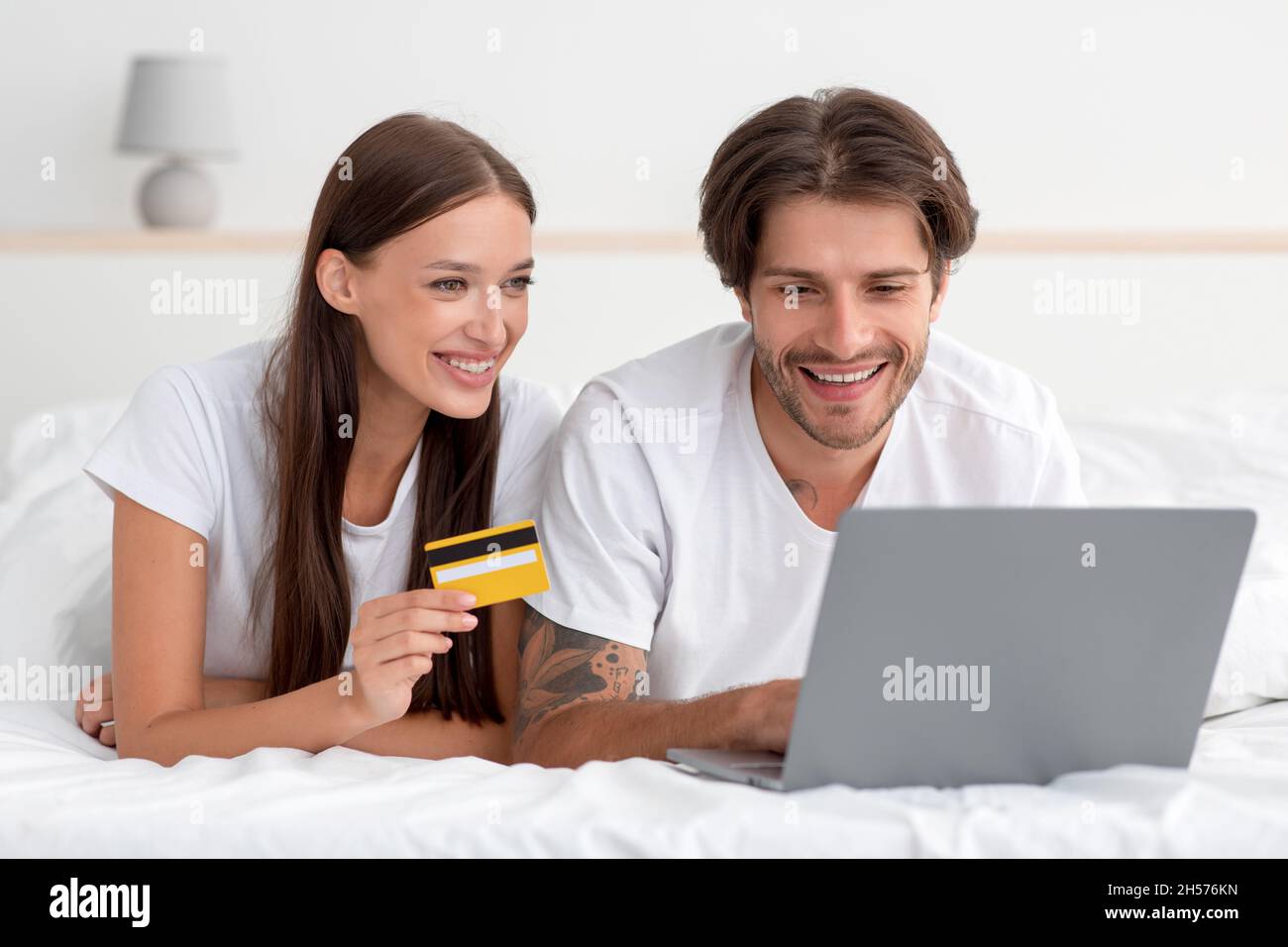 Satisfied millennial caucasian male and female lie on bed, paying for purchase with laptop and credit card in bedroom Stock Photo