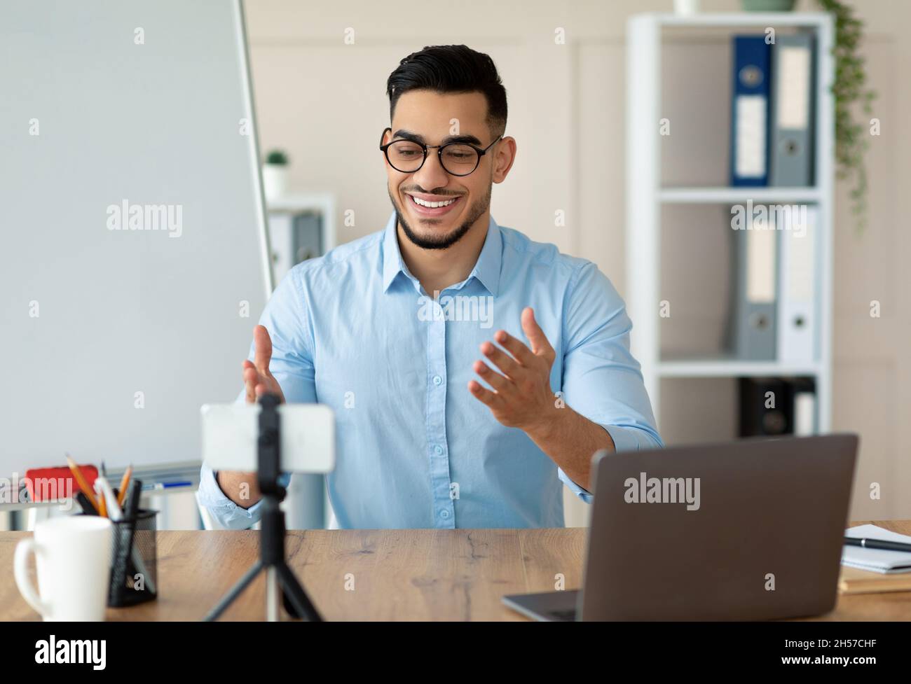 Happy Arab male teacher giving online lesson, using smartphone and laptop for remote education at home office Stock Photo