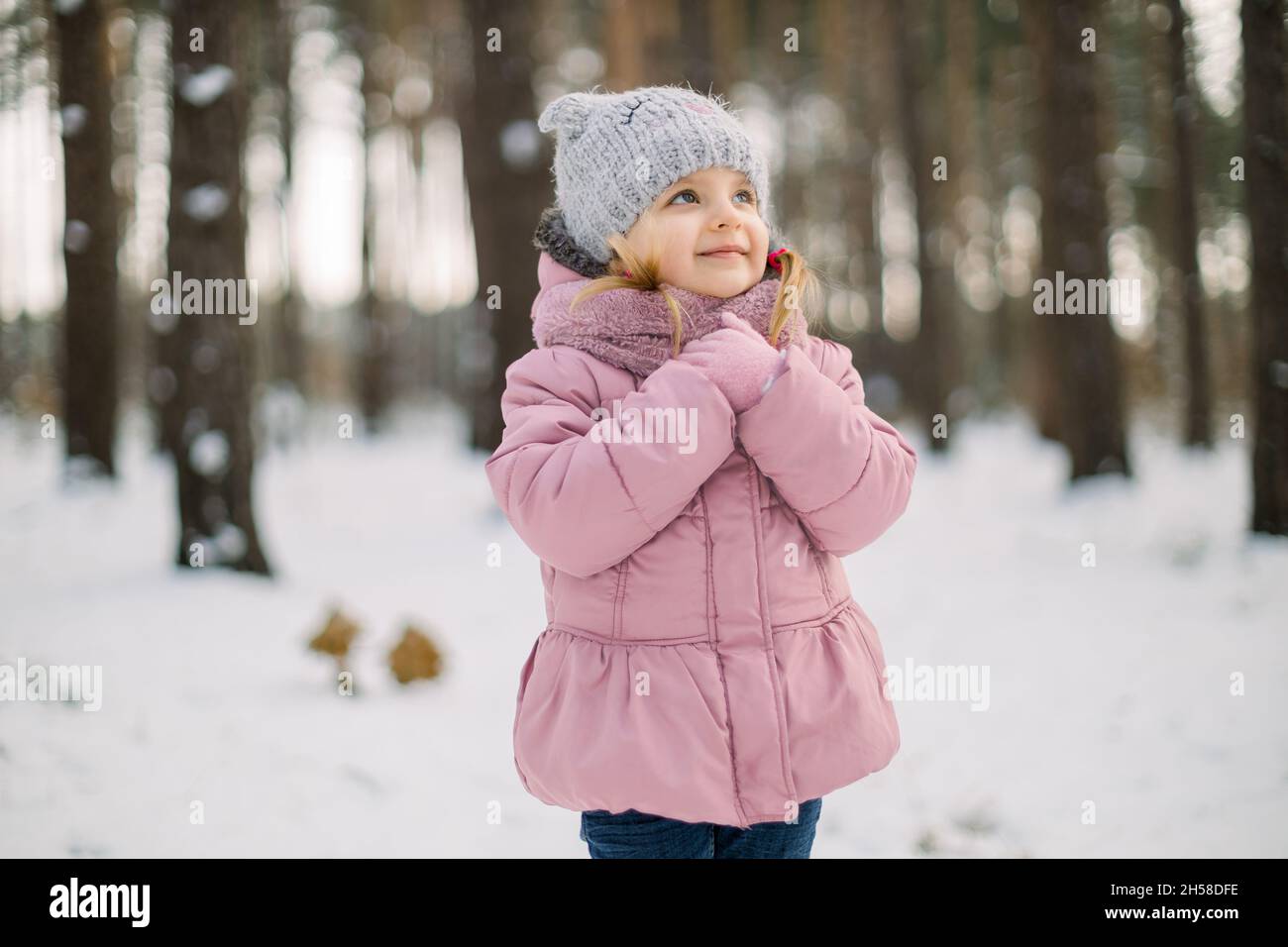 Dreamy cute little girl in stylish warm winter clothes, posing outdoors in  the snowy forest and looking up. Pretty child girl in pink outfit enjoying  winter season and snow in the nature