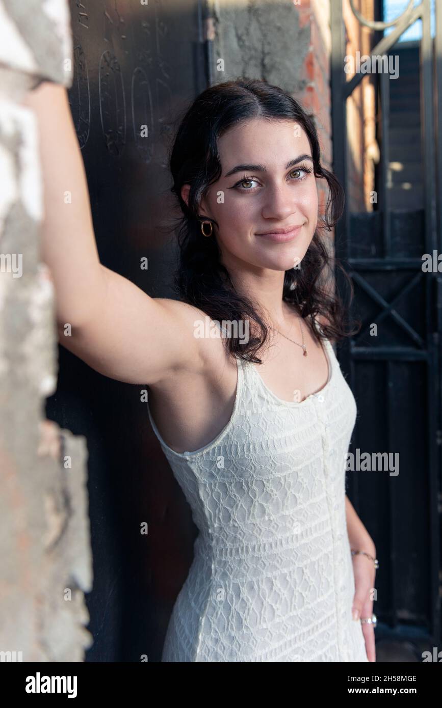 Confident high school senior girl spans her arms in rustic brick building doorway posing in her lace white dress. Stock Photo
