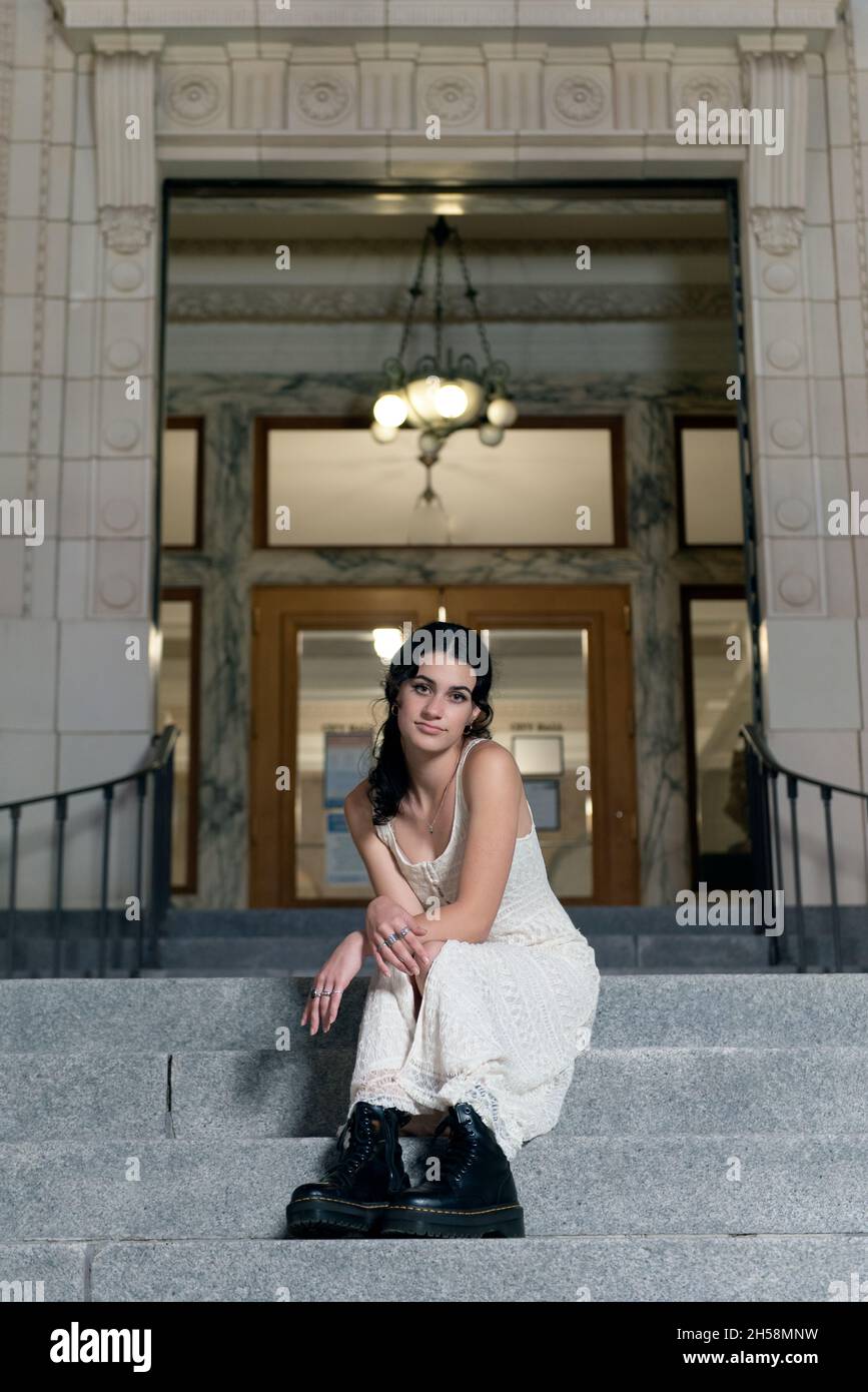 Confident high school senior girl sits on steps of elegant architecture building with lamps illuminated at dusk. Stock Photo