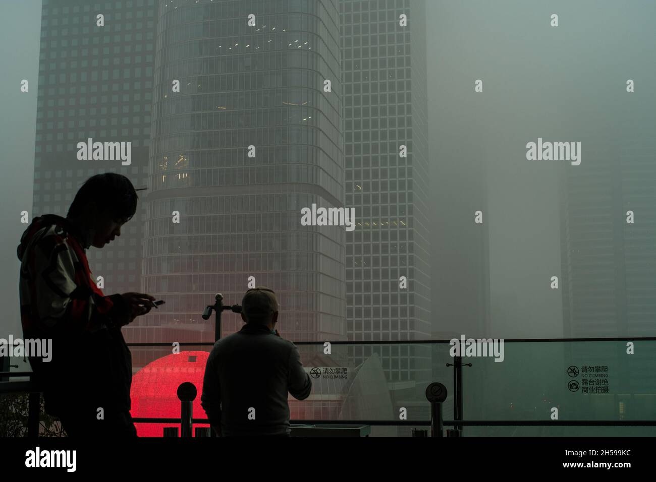 A young man checks his mobile phone while another looks at the skyscraper shrouded in severe smog in Beijing CBD, China. 06-Nov-2021 Stock Photo