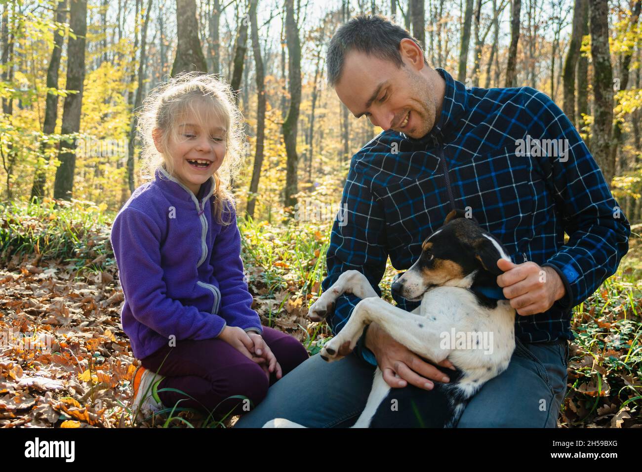A man in plaid coffee and jeans is stroking a dog lying on his lap. A girl is sitting next to him. People are smiling. Father and daughter in the autu Stock Photo
