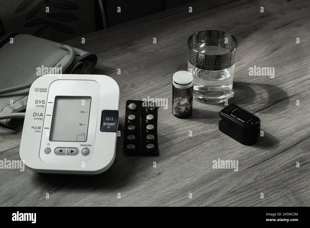 On a wooden table there is an automatic tonometer, a glass of water, pills, and an oxyometer. Black and white photo. Health Concept Stock Photo