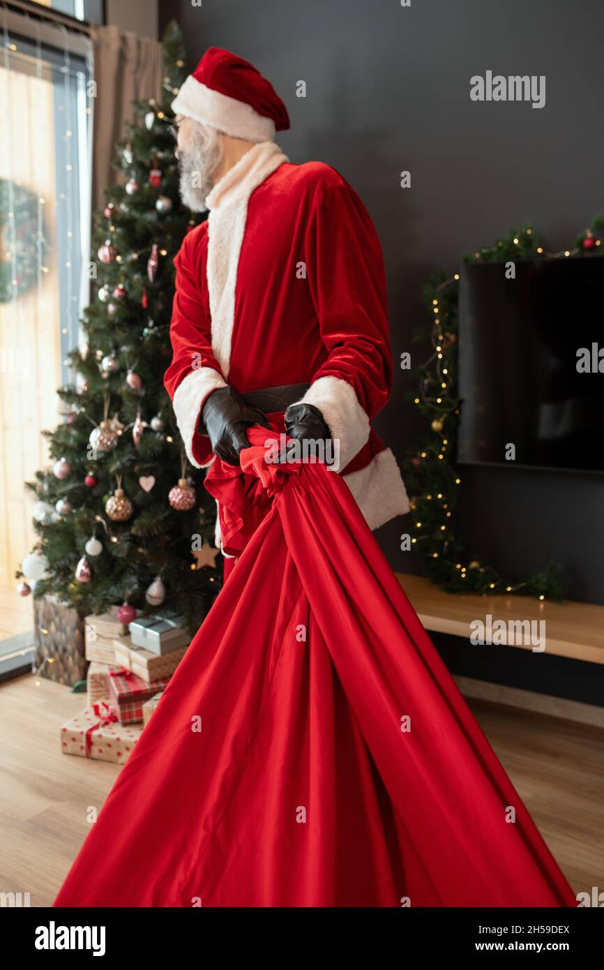 Santa Claus in red costume dragging big sack of Christmas gifts to decorated tree in cozy house Stock Photo