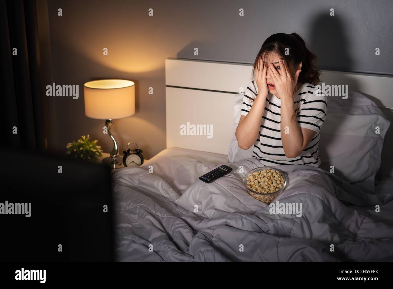scared young woman is watching horror movie TV on a bed at night Stock Photo