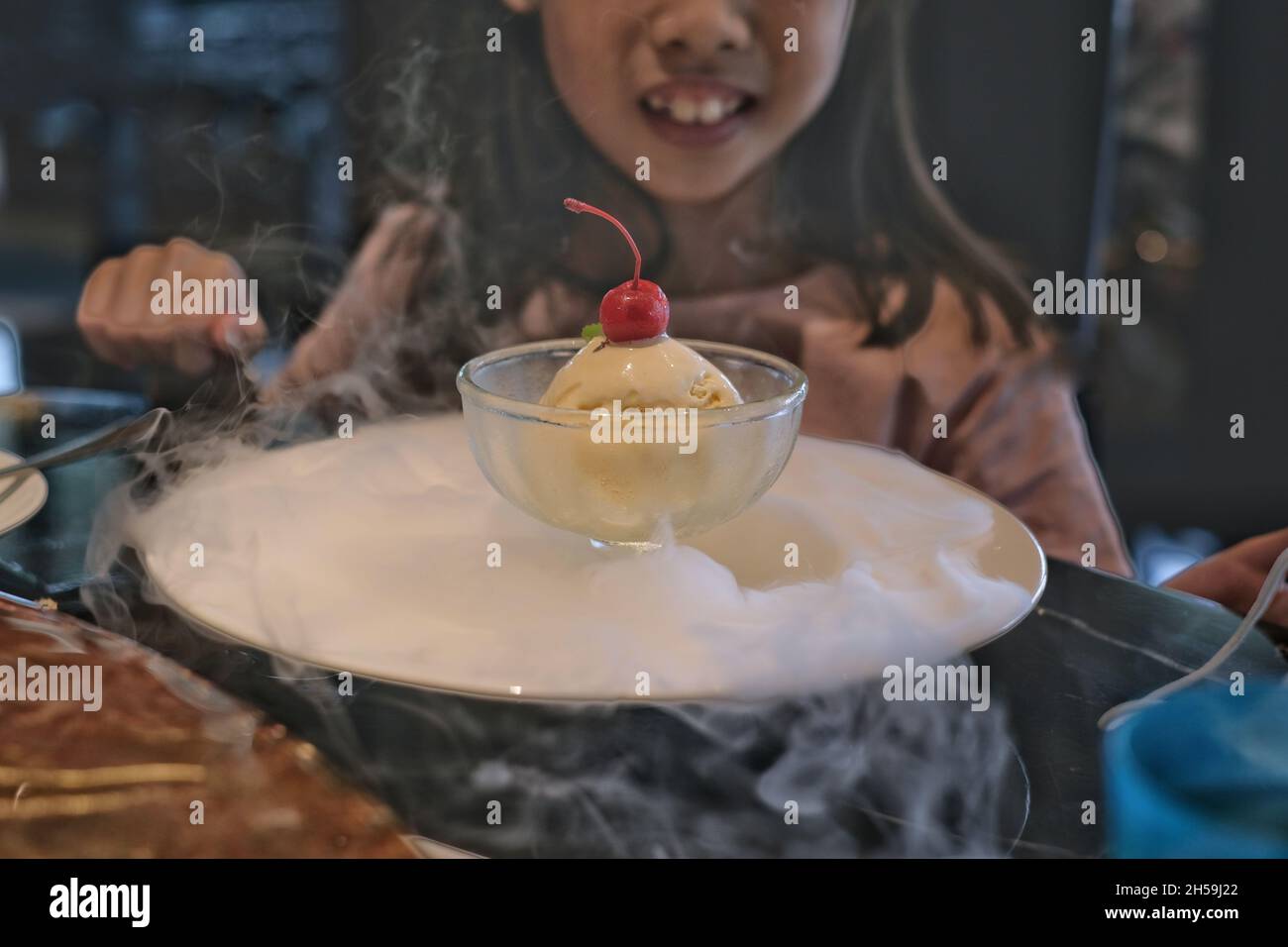 A cute young Asian girl is sitting in a restaurant, eating a bowl of vanilla ice cream, decorated by white smoke from dry ice, with cherry topping.  K Stock Photo