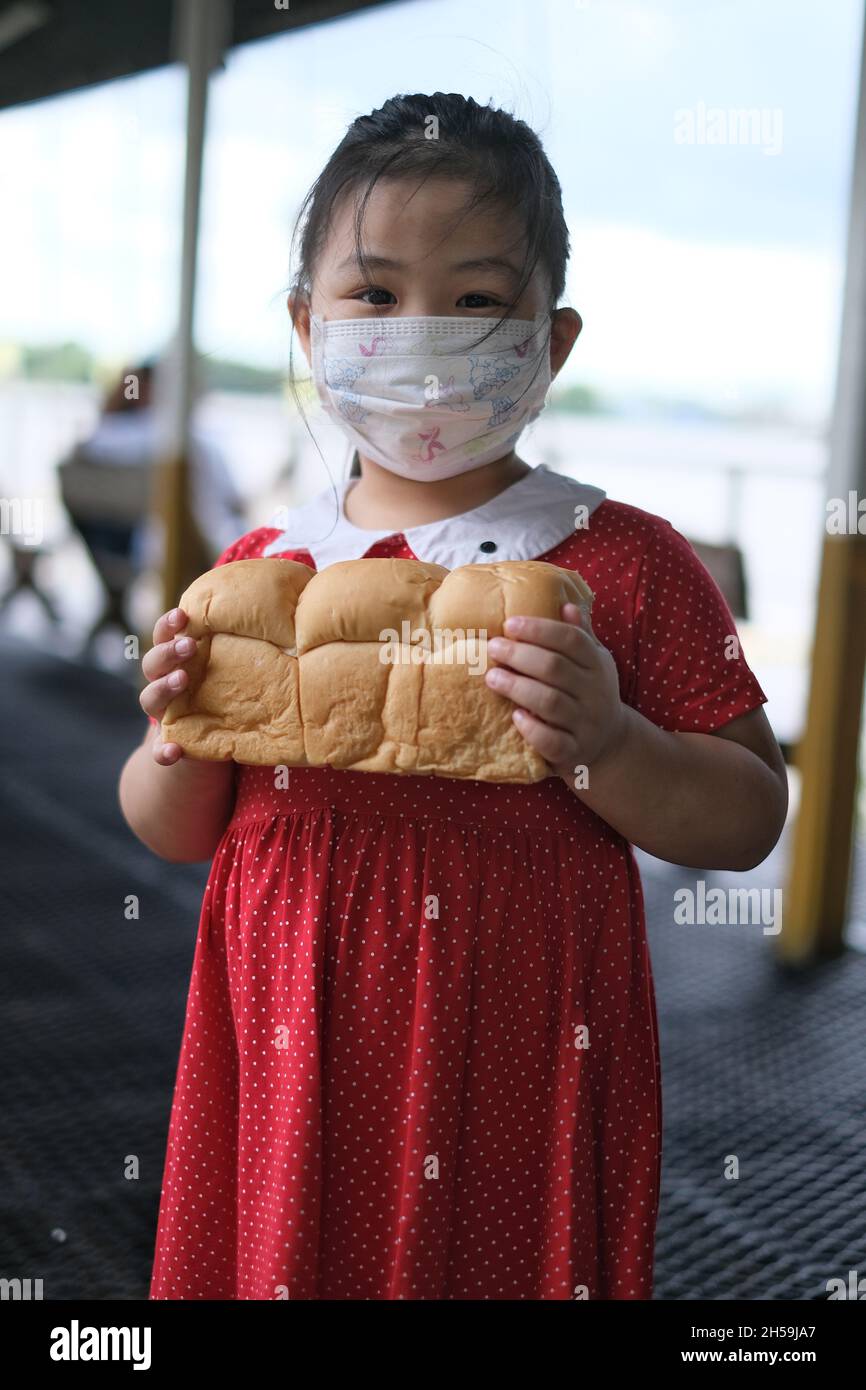 A cute young Asian girl in bright red dress with face mask is hold a large loaf of soft brown bread. Stock Photo