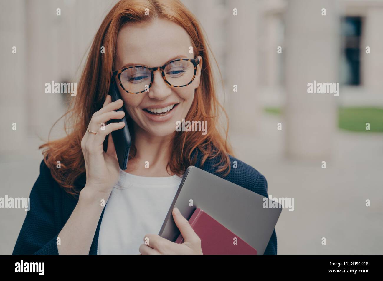 Pleased red-haired businesswoman with laptop discussing something with partner via mobile phone Stock Photo