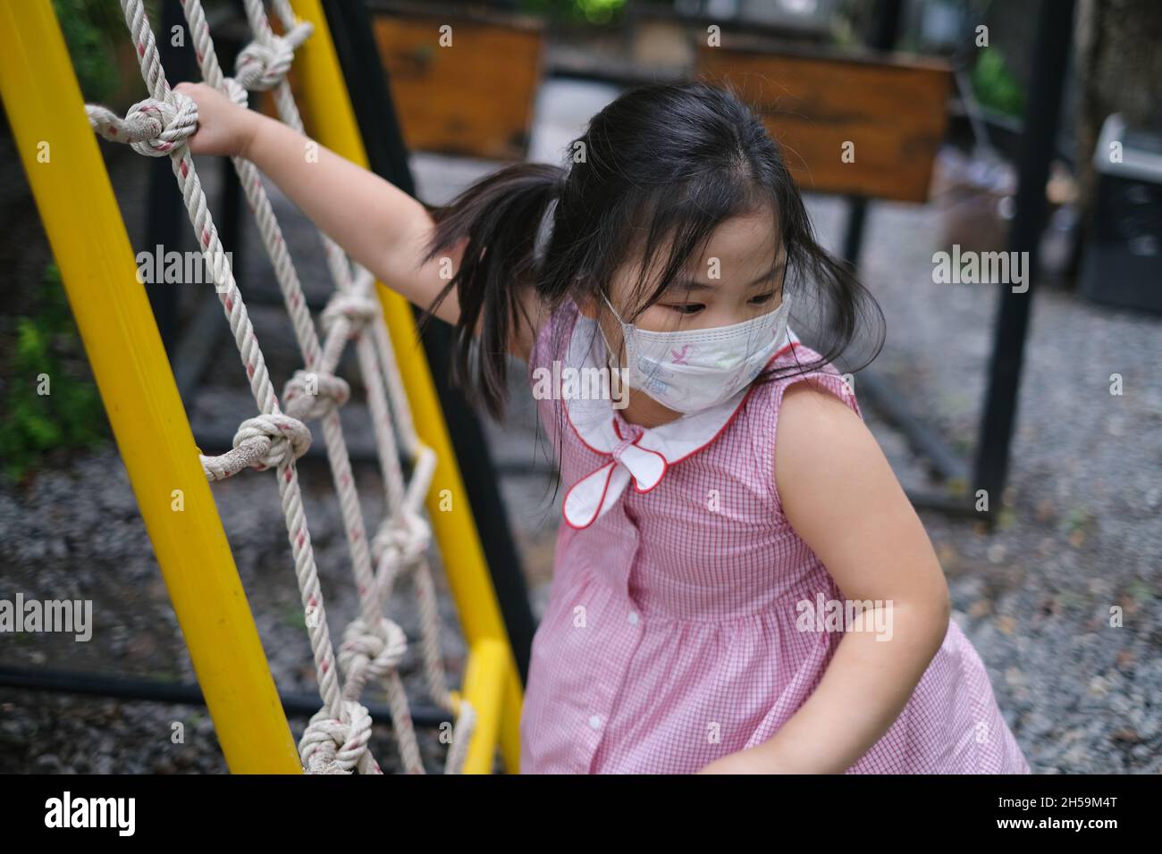 A cute young Asian girl, with white face mask, is playing alone on a jungle gym in a playground, climbing a ramp and nets made from rope during Covid- Stock Photo