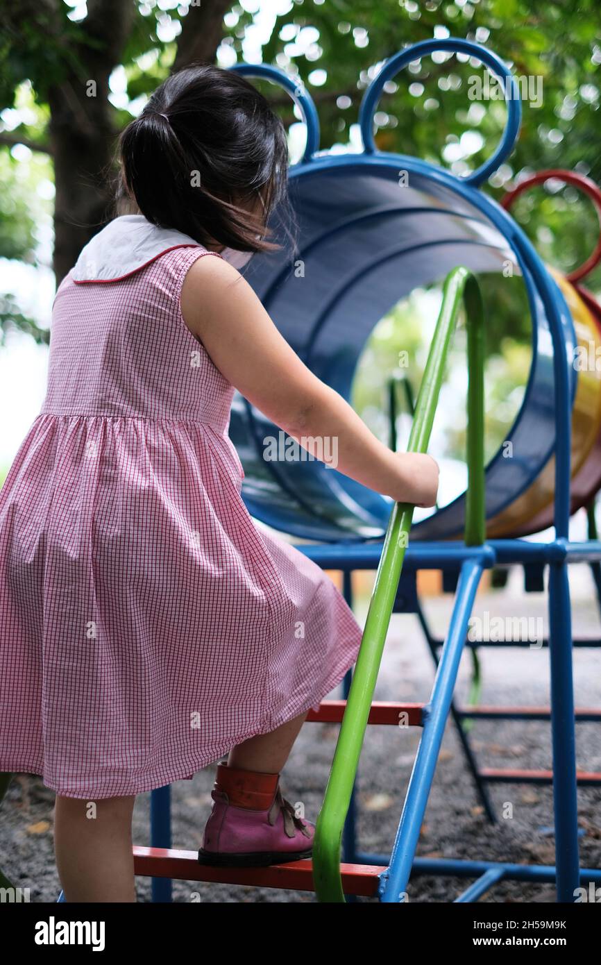 A cute young Asian girl, with white face mask, is playing alone on a jungle gym in a playground, climbing a ramp and nets made from rope during Covid- Stock Photo