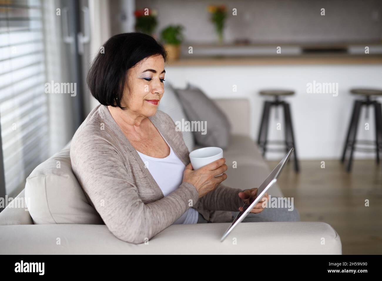 Happy senior woman with tablet sitting on sofa indoors at home, video call concept. Stock Photo