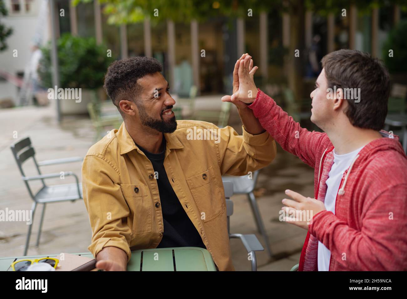 Young man with Down syndrome with mentoring friend sitting outdoors in cafe celebrating success. Stock Photo