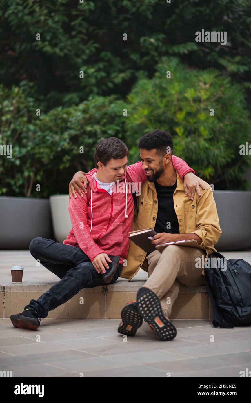 Happy young man with Down syndrome and mentoring friend sitting with arms around outdoors Stock Photo