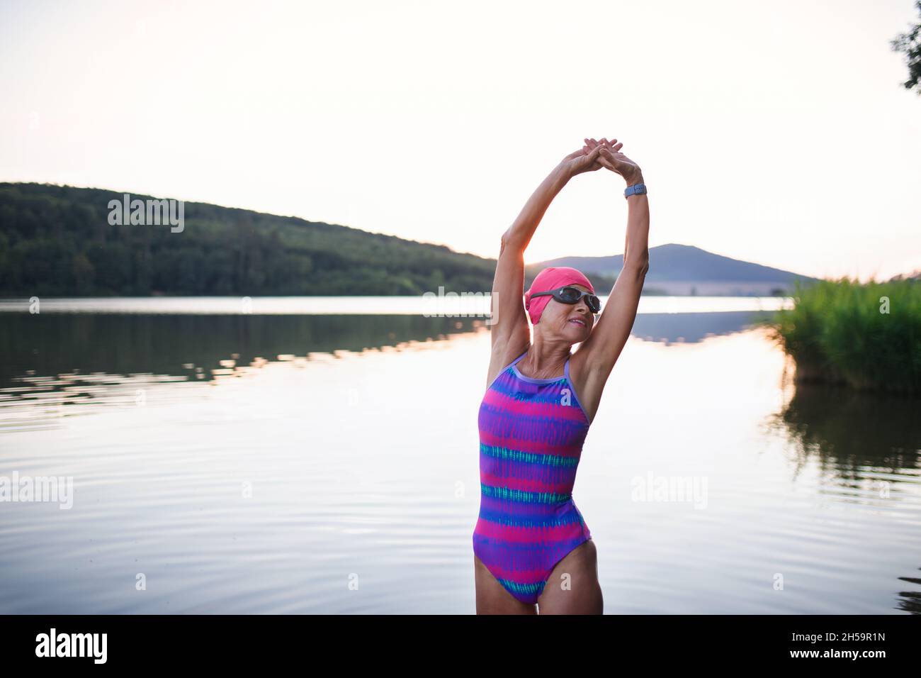 Portrait of active senior woman swimmer standing and stretching outdoors by lake. Stock Photo